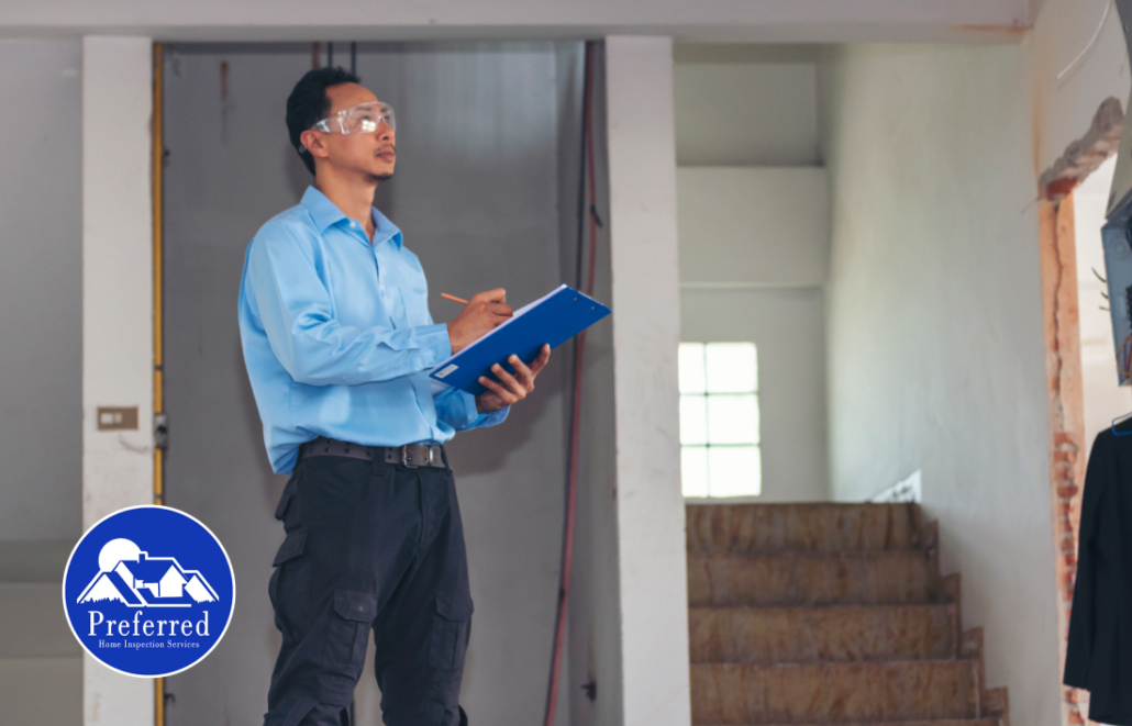 An adult man with dark hair stands inside a building under construction. He is wearing safety glasses, a blue button-up shirt, and dark work pants with multiple pockets. Holding a blue clipboard and pencil, he looks to the side while inspecting the site. Sunlight streams through a nearby window, illuminating the unfinished interior and staircase. This image is ideal for representing construction, engineering, building inspections, workplace safety, and industrial projects.